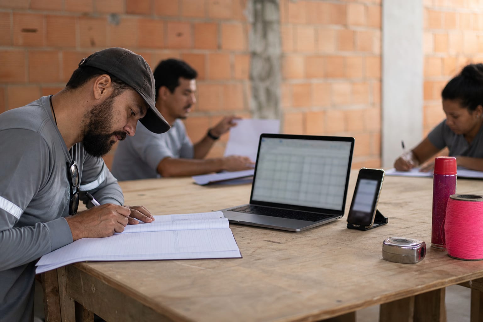 Livro de obra impresso, notebook e celular lado a lado sobre bancada de compensado no escritório de uma obra
